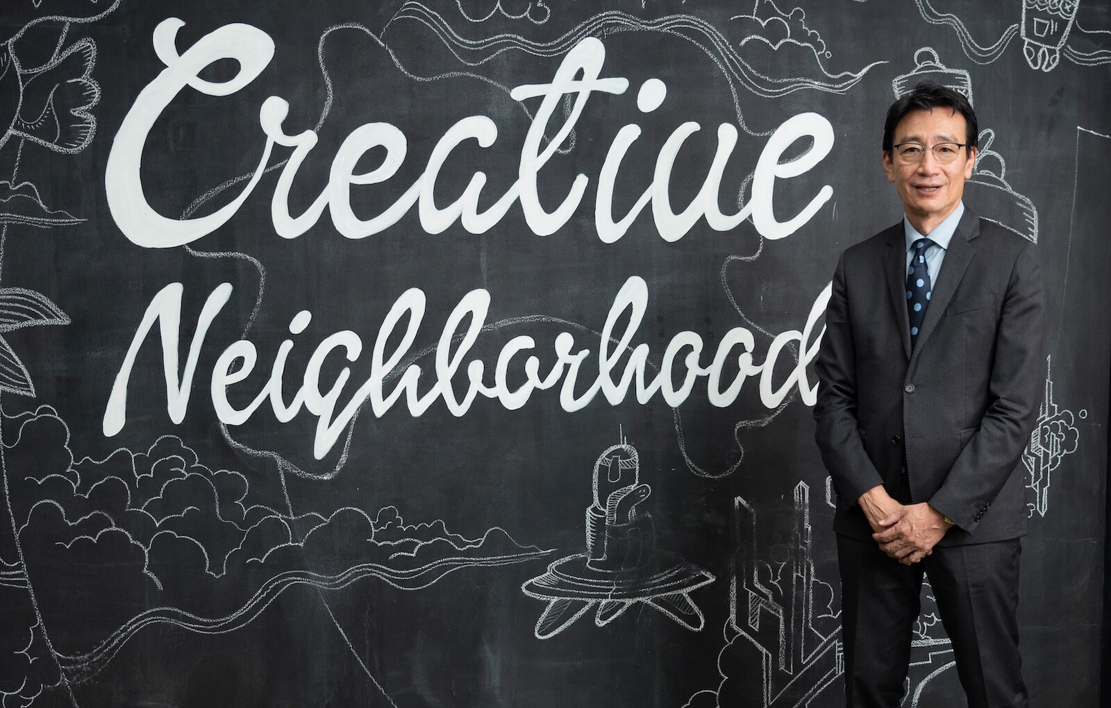 黒板の前にスーツ姿の男性が立っている。背景の黒板には大きく英語で「Creative Neighborhood」と白い文字が書かれ、周囲に街や建物をイメージしたチョークのイラストが描かれている。