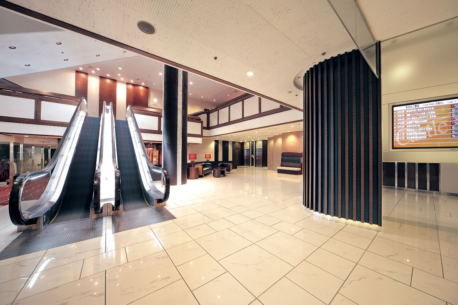 ホテルのロビー空間。吹き抜けの広いフロアにエスカレーターが設置され、光沢のある床と間接照明が上質な雰囲気を演出している。奥にはソファが並ぶラウンジスペースがあり、壁面には案内表示のデジタルサイネージが設置されている。