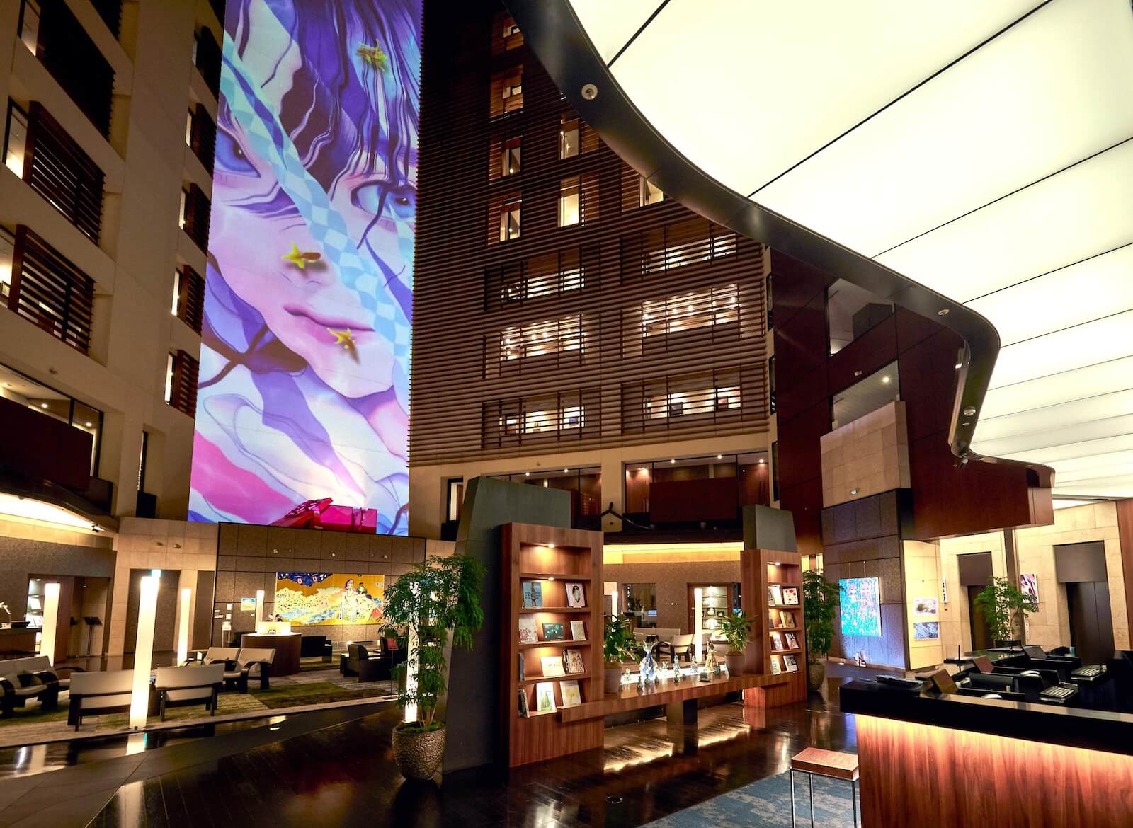 25階アトリウムのロビー全景。壁面いっぱいに女性の顔を描いた大型アートが投影され、書棚や展示台が配置された空間全体が巨大なギャラリーのように構成されている。