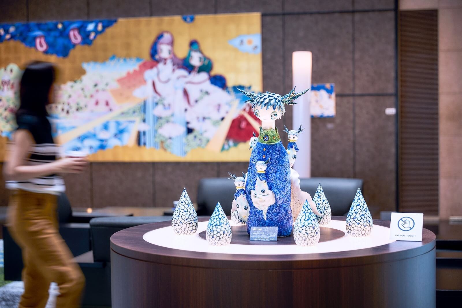 25階アトリウムに展示された立体作品。円形の展示台中央に青を基調としたオブジェが置かれ、背後の壁面には大型の絵画が掛けられており、空間全体がギャラリーのように演出されている。