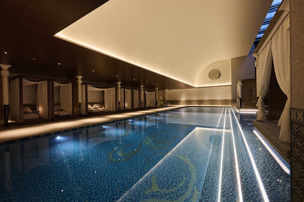 天井の高い屋内プール。青いモザイクタイルの水面と、カーテン付きのデイベッドが並ぶ落ち着いた空間。