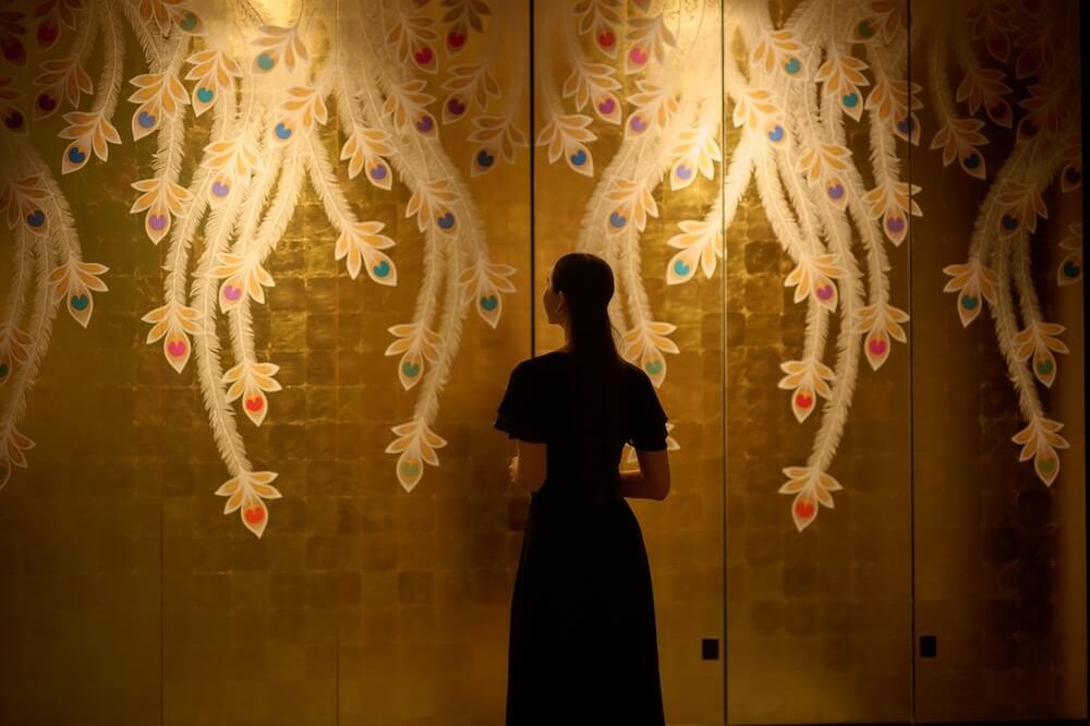 金色の壁一面に、羽のような模様が描かれている。手前に黒い服を着た人物が立ち、壁の装飾を見上げている。