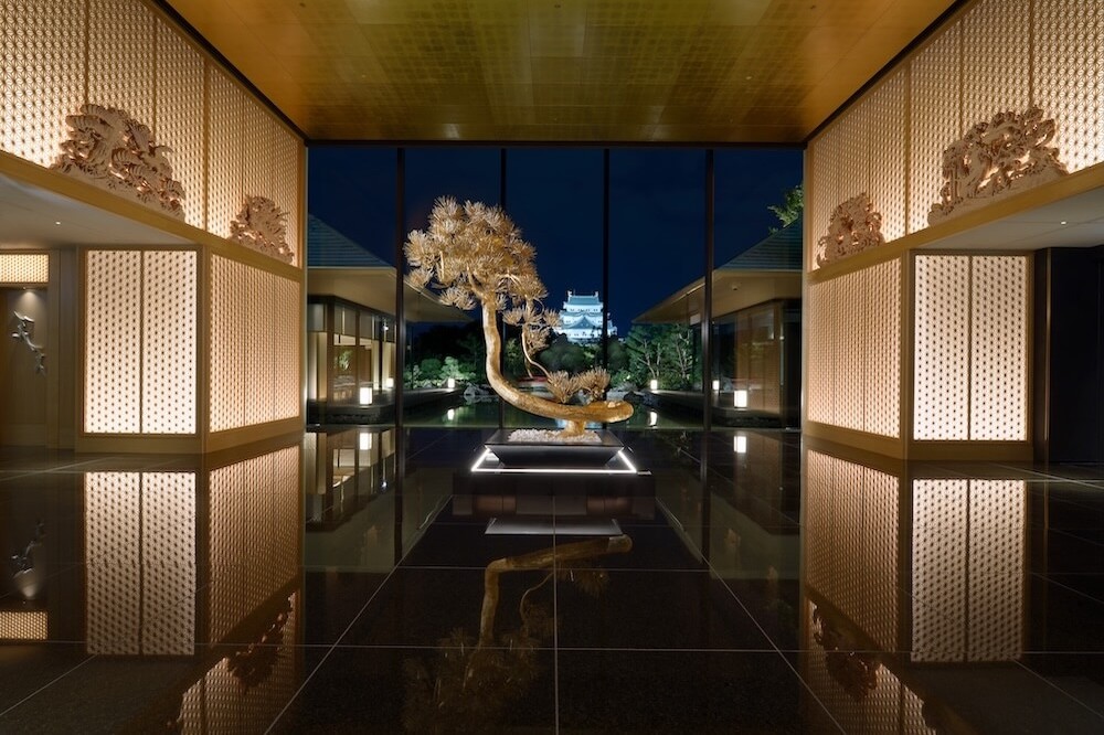 夜の屋内通路。黒く光る床の中央に台座があり、盆栽のような樹木が照明に照らされて置かれている。正面の大きな窓の外に城の建物が見える。