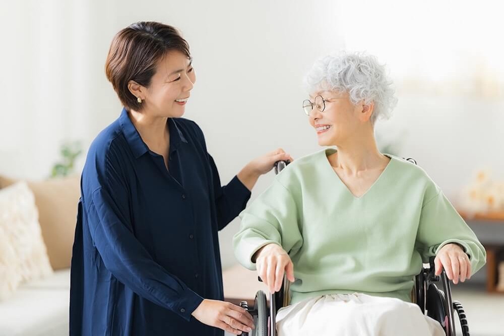 高齢の女性が車いすに座り、隣に立つ女性が笑顔で手を添えて会話している室内の様子。