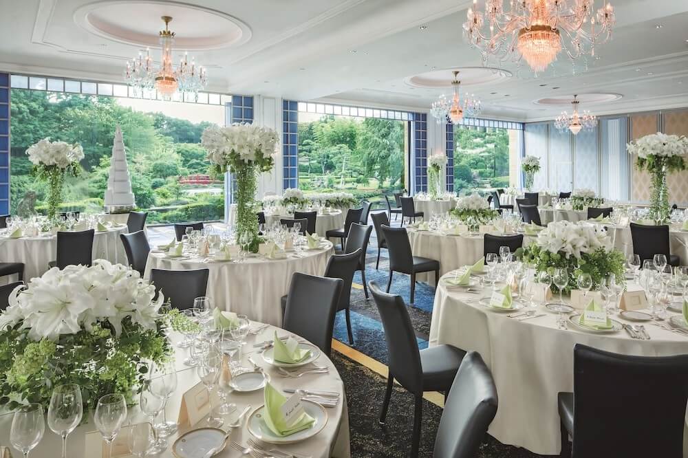 大きな窓から緑の庭園が見える明るい宴会場。白い円卓が並び、白とグリーンの花を高くあしらった装花、グラスとカトラリーが整えられている。天井にはシャンデリアが下がり、結婚披露宴の準備が整った室内。