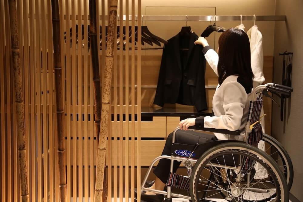 車椅子に座った人物が、木製のクローゼットで上着をハンガーに掛けている。低い位置に衣類が配置されている。