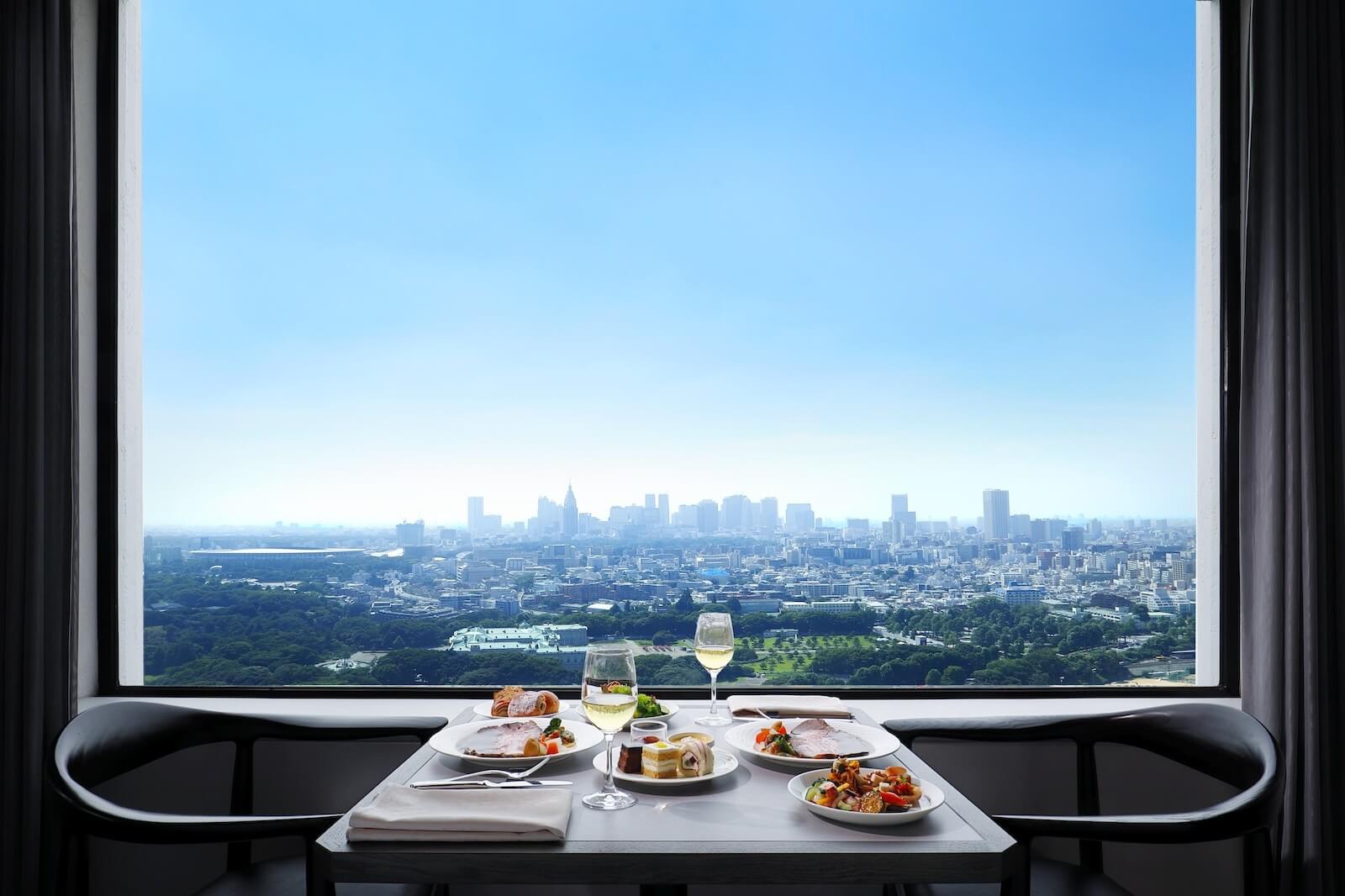 大きな窓の前にテーブルが置かれ、料理とワイングラスが並んでいる。窓の外には青空の下、街並みと広い緑地が一望できる。