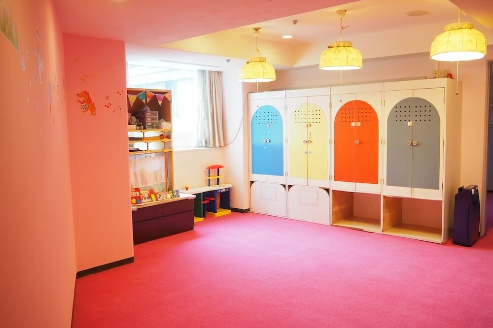 ピンク色の床の明るい部屋。おもちゃ棚と小さな机があり、壁一面に青・黄・オレンジ・グレーのロッカーが並んでいる。