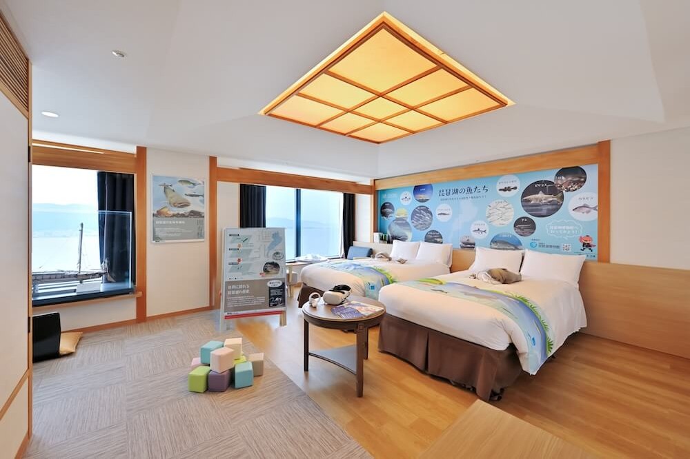 びわ湖に面した窓のある客室。和モダンの内装で、ツインベッドのヘッドボードには海や魚をモチーフにしたイラストと解説パネルが設えられている。室内には木製家具と畳スペースがあり、子ども向けの積み木や展示スタンドも配置された体験型の客室。