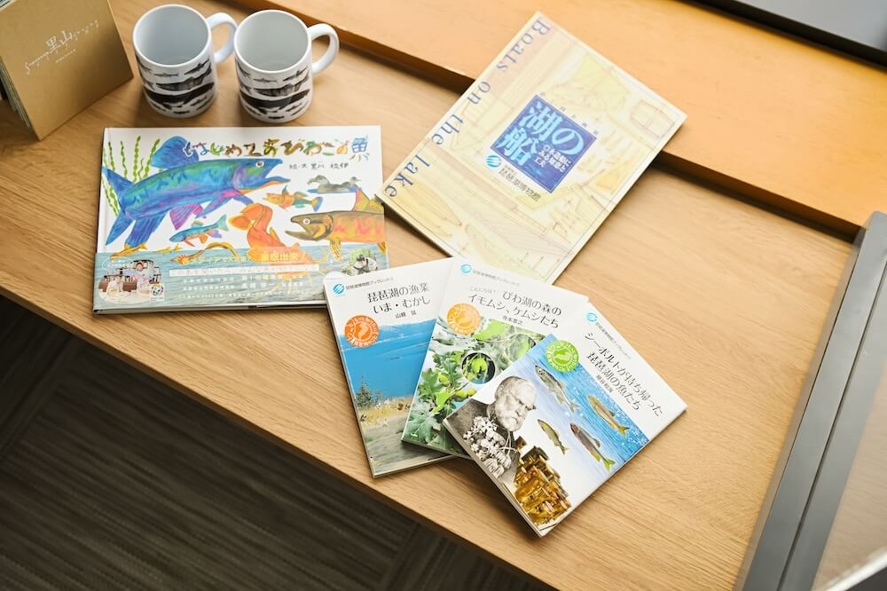 客室のデスク上に並べられた、地域の自然や海の生き物をテーマにした図鑑などの冊子。魚のイラストが描かれた表紙の、大きさが違う冊子が置かれ、マグカップも添えられている。びわ湖について学べる滞在用アイテム。