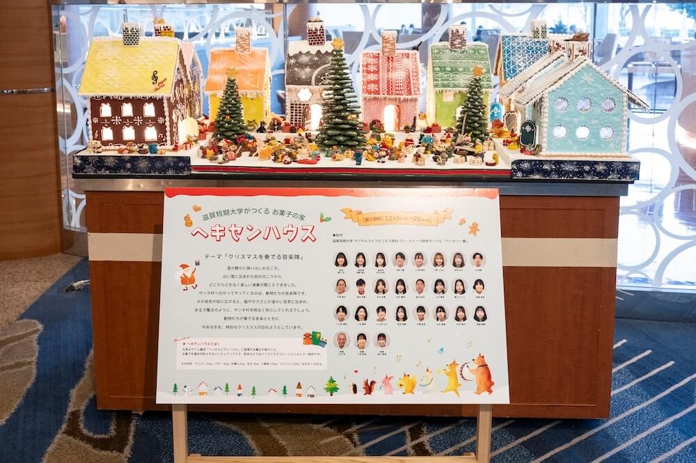 滋賀短期大学のパン・スイーツ研究サークル学生によるヘキセンハウス展示と紹介パネル。制作テーマや参加学生の顔写真を掲示し、学生が主体的に活躍できる場を創出していることを伝える展示。