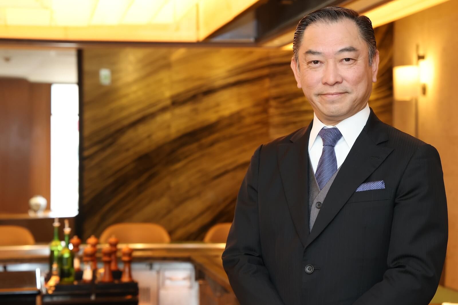 スーツ姿の男性が、落ち着いた照明のレストラン内に立っている。背後には木目調の壁とカウンター席、テーブル上に調味器具が見える。
