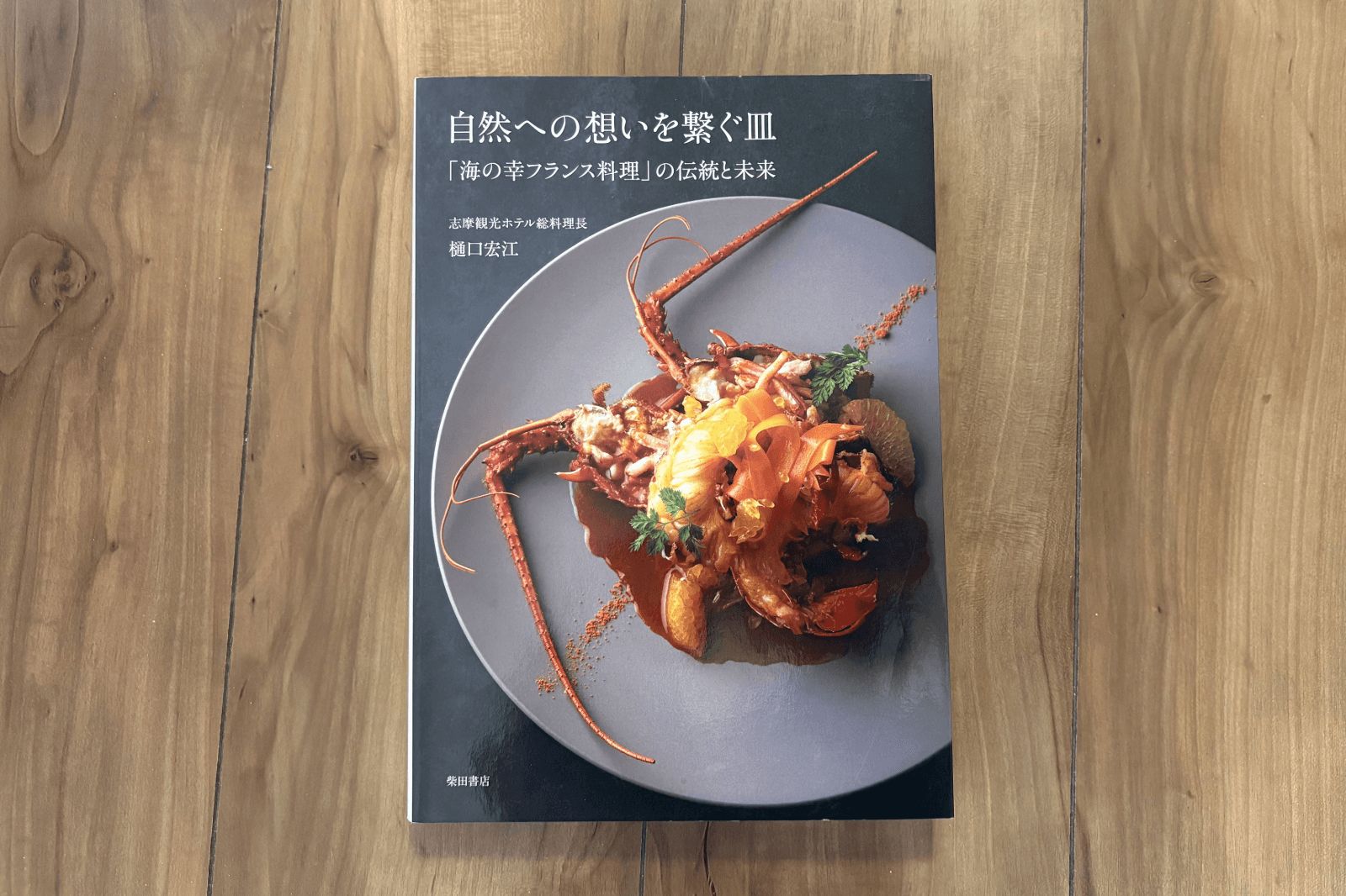 木目の床に置かれた料理本の表紙。黒地に伊勢海老料理の写真と「自然への想いを繋ぐ皿」「『海の幸フランス料理』の伝統と未来」「志摩観光ホテル総料理長 樋口宏江」「柴田書店」の文字が記されている。