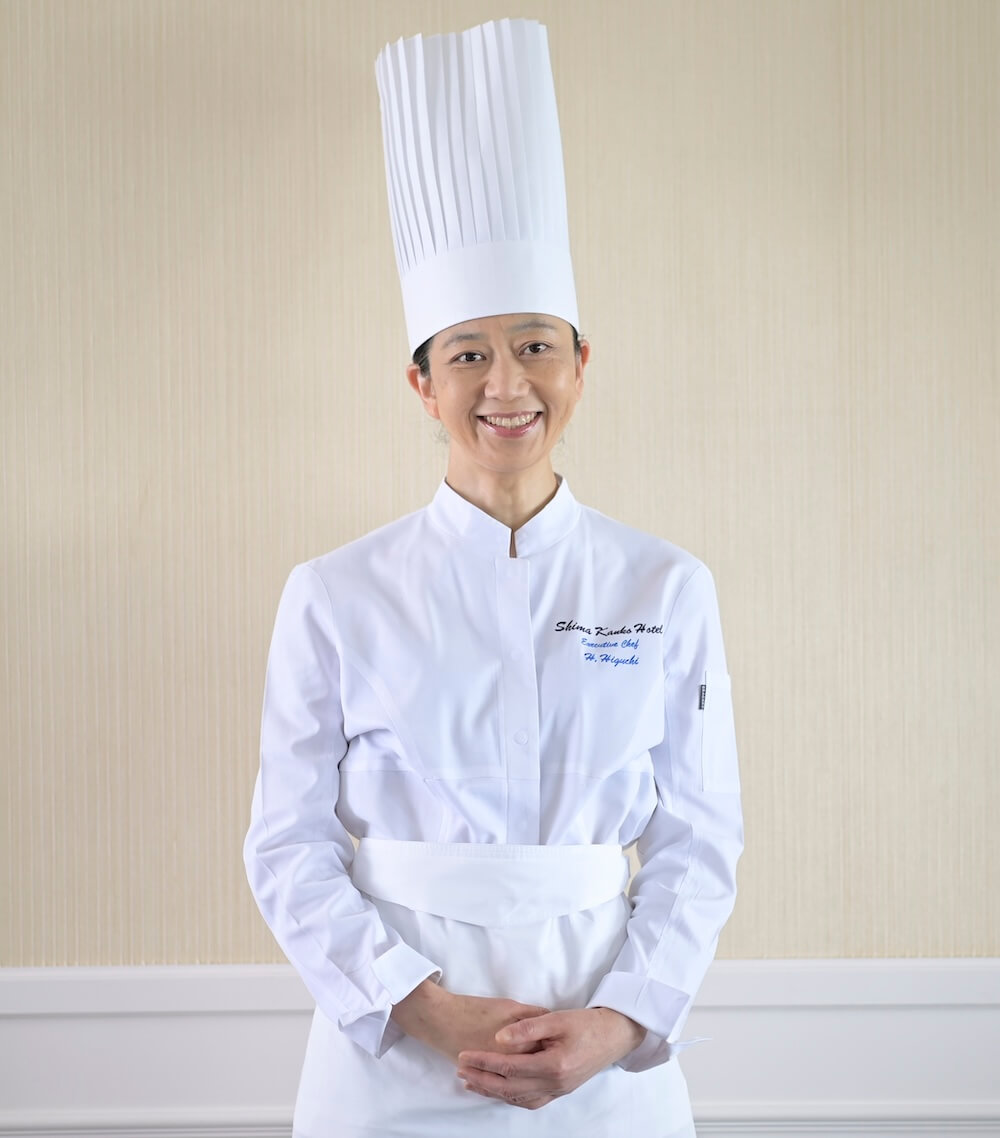 白いコックコートとコック帽を着用し、正面を向いて微笑む立ち姿。志摩観光ホテル総料理長の樋口宏江シェフ。