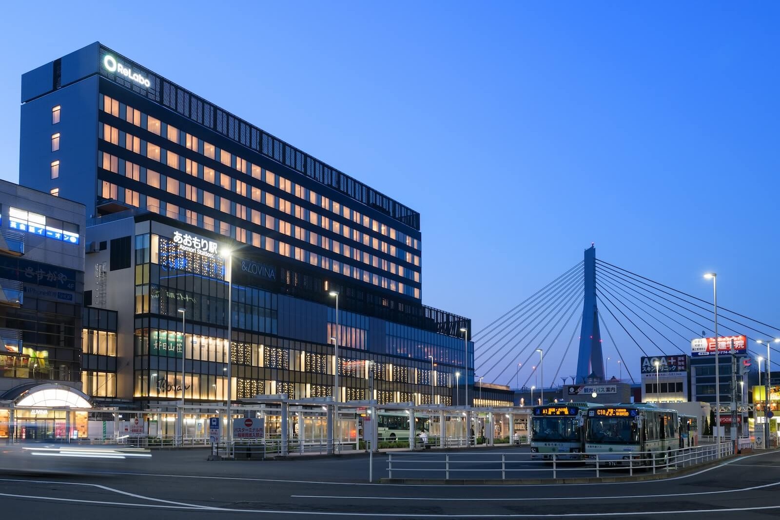 夕暮れ時または明け方の青森駅前。駅前広場とバス乗り場の奥に、駅に直結したホテルの大きな建物が立ち、客室の窓明かりが横一列に灯っている。右奥には斜張橋が空に浮かんで見える。