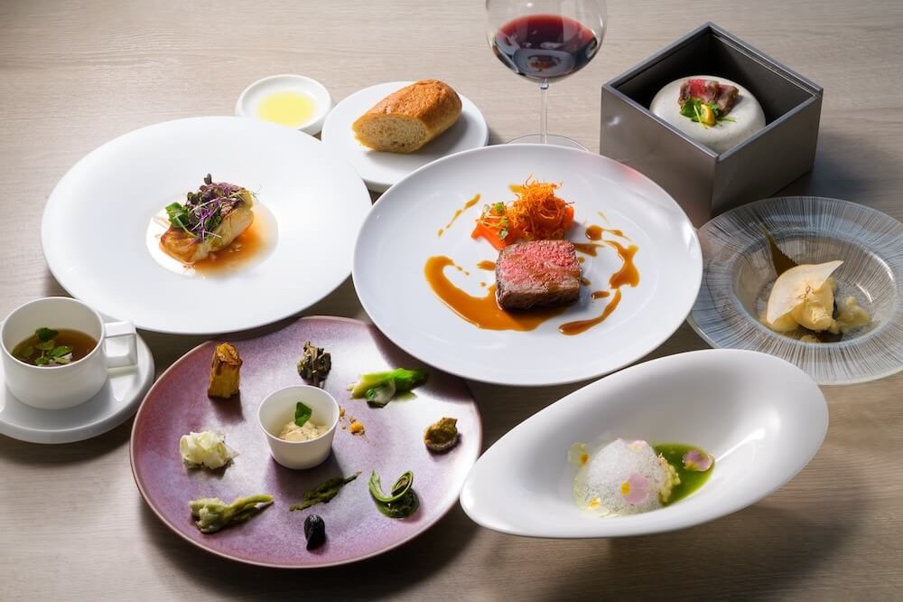 テーブルに並べられたコース料理。肉料理を中心に、前菜、スープ、パン、デザートが白い器に盛られ、横に赤ワインのグラスが置かれている。