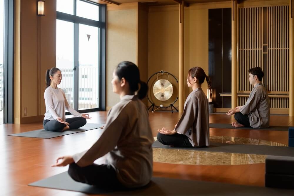 木の床の和モダンな室内で、4人がヨガマットの上に座り、目を閉じて瞑想している。窓からやわらかな光が入り、奥には大きな銅鑼が置かれている。