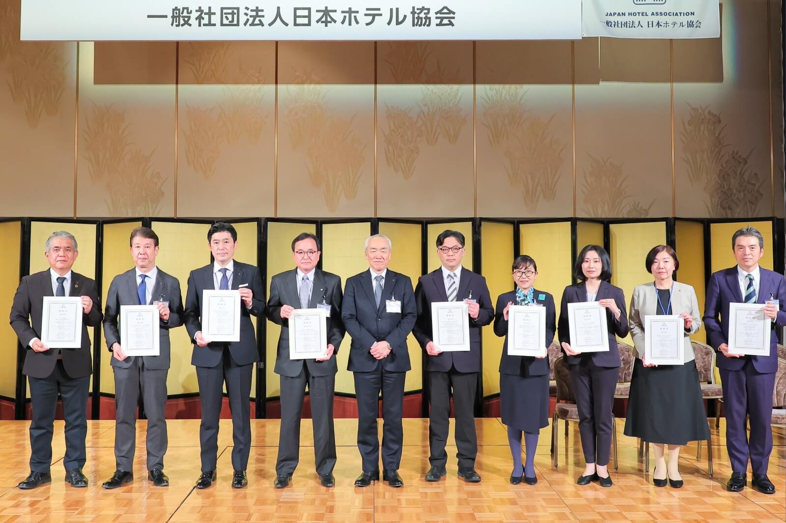 一般社団法人日本ホテル協会主催の第7回「会員ホテルの社会的貢献に対する会長表彰」の会場(リーガロイヤルホテル大阪 ヴィニェット コレクション)で、表彰状を手に並ぶ受賞者と関係者の集合写真。壇上で複数名が整列している。