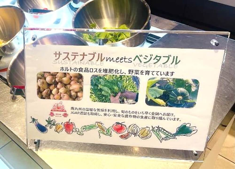 「サステナブル meets ベジタブル」と書かれた案内パネルがバイキング会場に設置されている様子。食品ロスを堆肥化して野菜を育てる取り組みを紹介し、ジャガイモや葉物野菜などの写真とイラストが掲載されている。