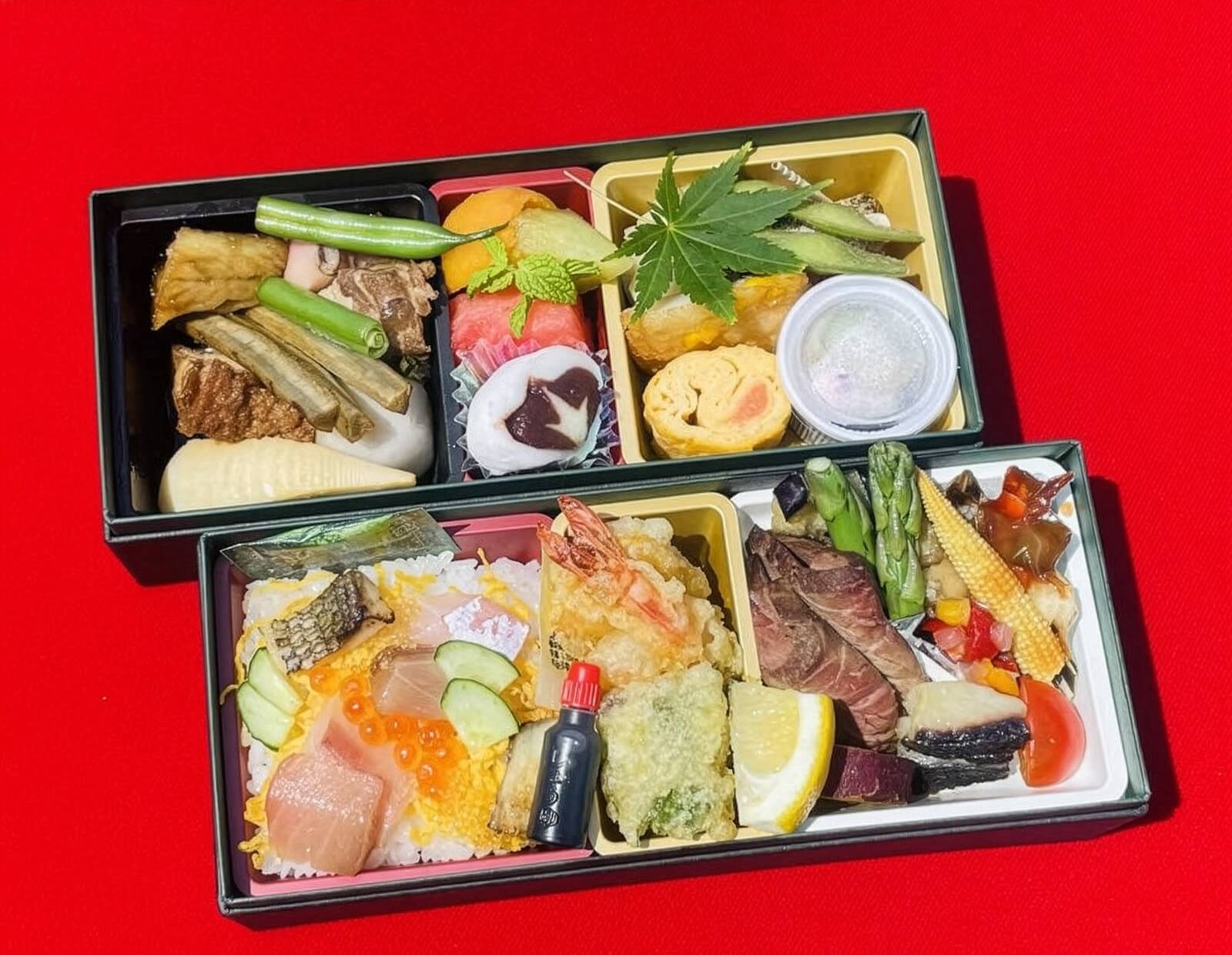 赤い背景の上に置かれた二段重の弁当。刺身やいくらが乗ったご飯、天ぷら、肉料理、煮物、卵焼き、野菜料理、フルーツなどが彩り豊かに詰められている。