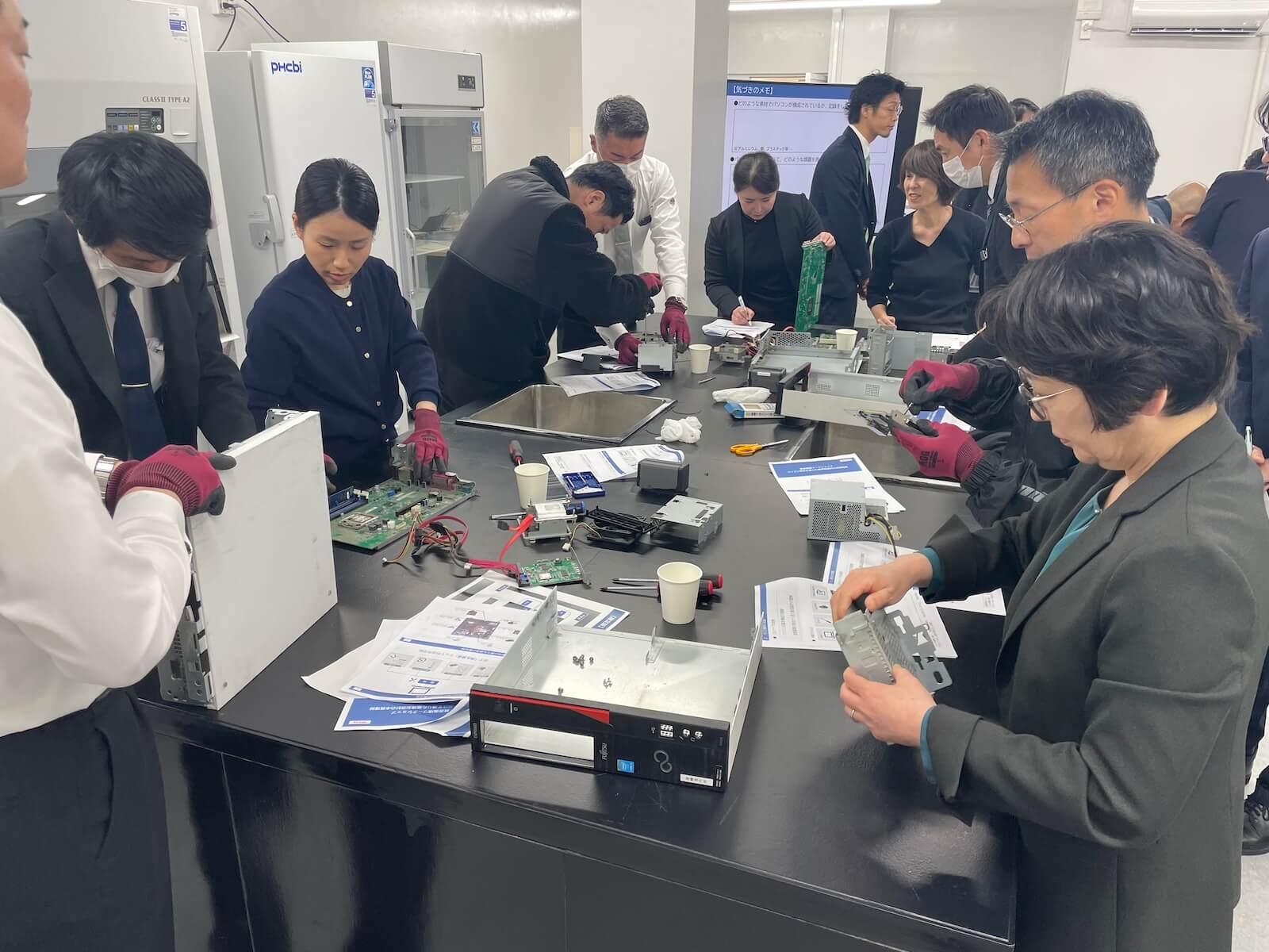 複数の参加者がテーブルを囲み、手袋を着用してパソコンを分解・分別している作業風景。基板や部品、工具、資料が並び、実習形式で取り組んでいる様子。