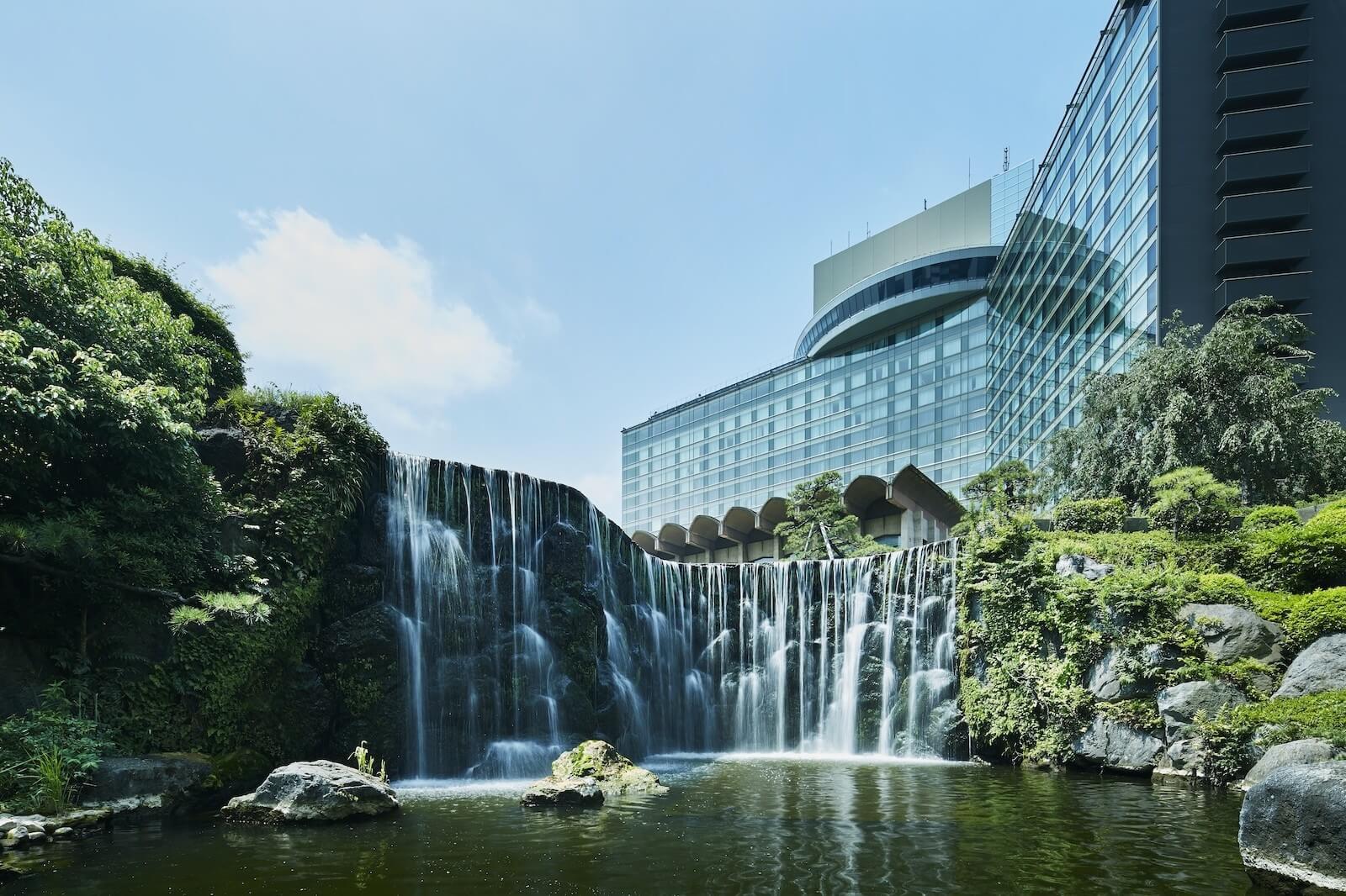 庭園の滝が流れ落ちる池の向こうに、大型ホテルの建物がそびえる景観。緑に囲まれた落ち着いた外観。