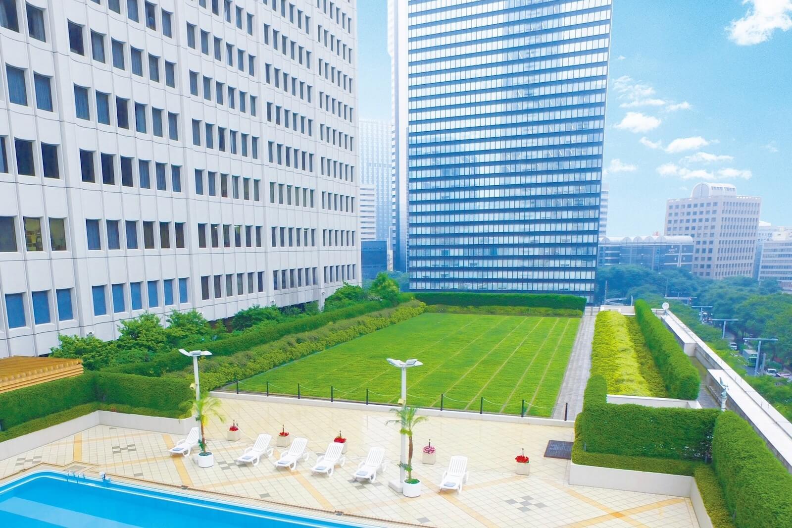 都市の高層ビルに囲まれたホテル屋外施設の芝生広場とプールサイドの様子。
