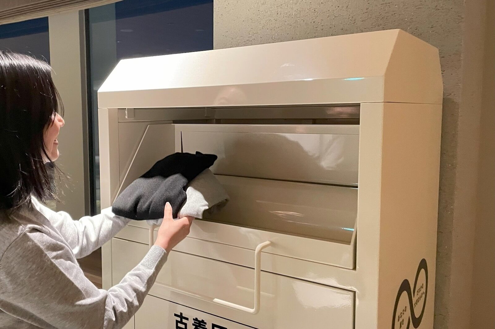 古着回収ボックスにセーターなどを入れる女性の様子。