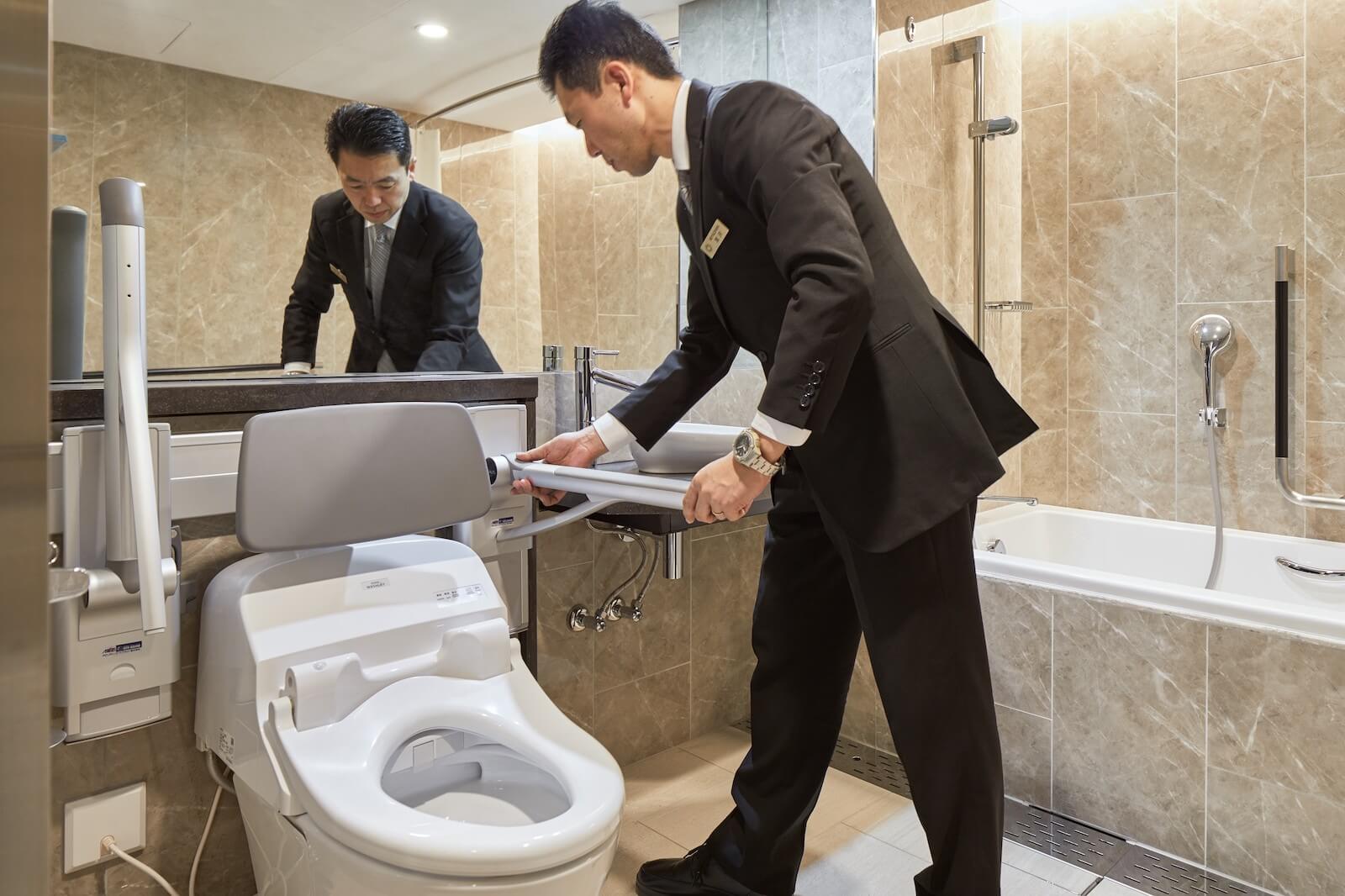 バリアフリー対応の浴室で、スタッフが可動式の手すり付きトイレ設備を調整している様子。