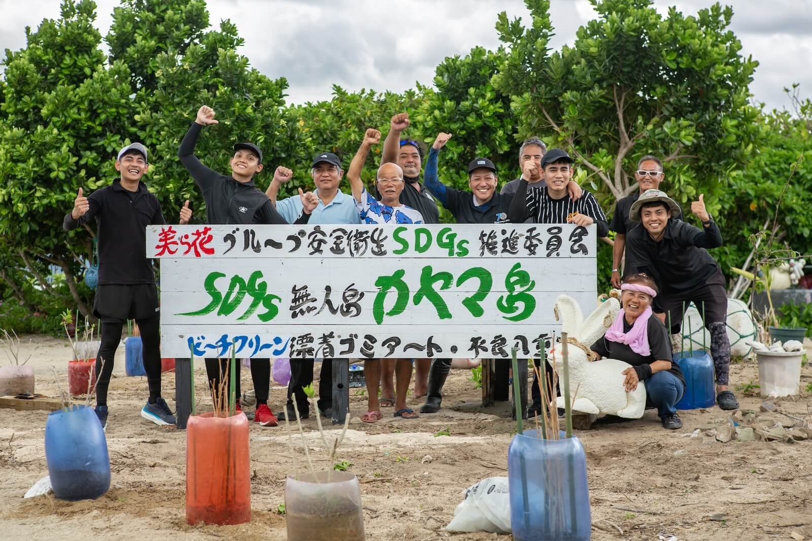 「SDGs 無人島カヤマ島」と書かれた看板の前で、ビーチクリーン活動に参加した人々がポーズをとっている様子。