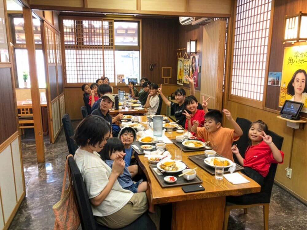 和風の店内で長いテーブルを囲み、食事を楽しみながらポーズを取る子どもたちと大人のグループ。