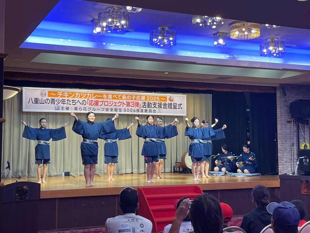 ステージ上で伝統衣装を着た子どもたちが踊りを披露し、観客が見守るイベントの様子。