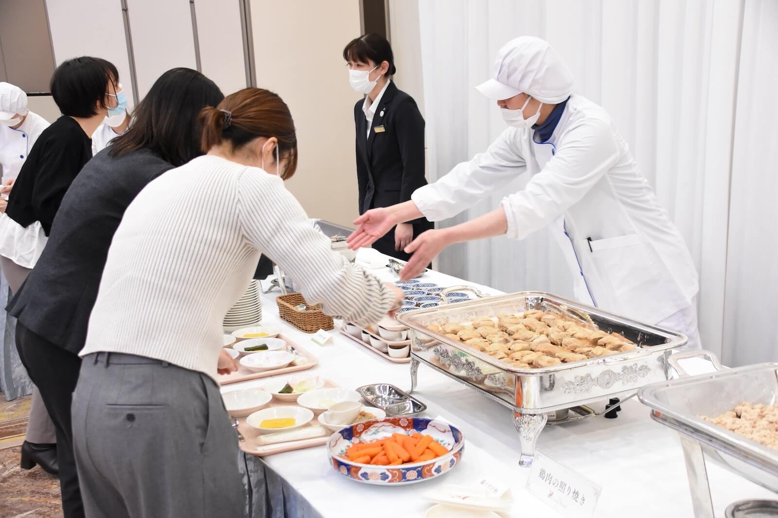 ビュッフェ形式の試食会でスタッフが料理を提供し、参加者がトレーに取り分けている様子。