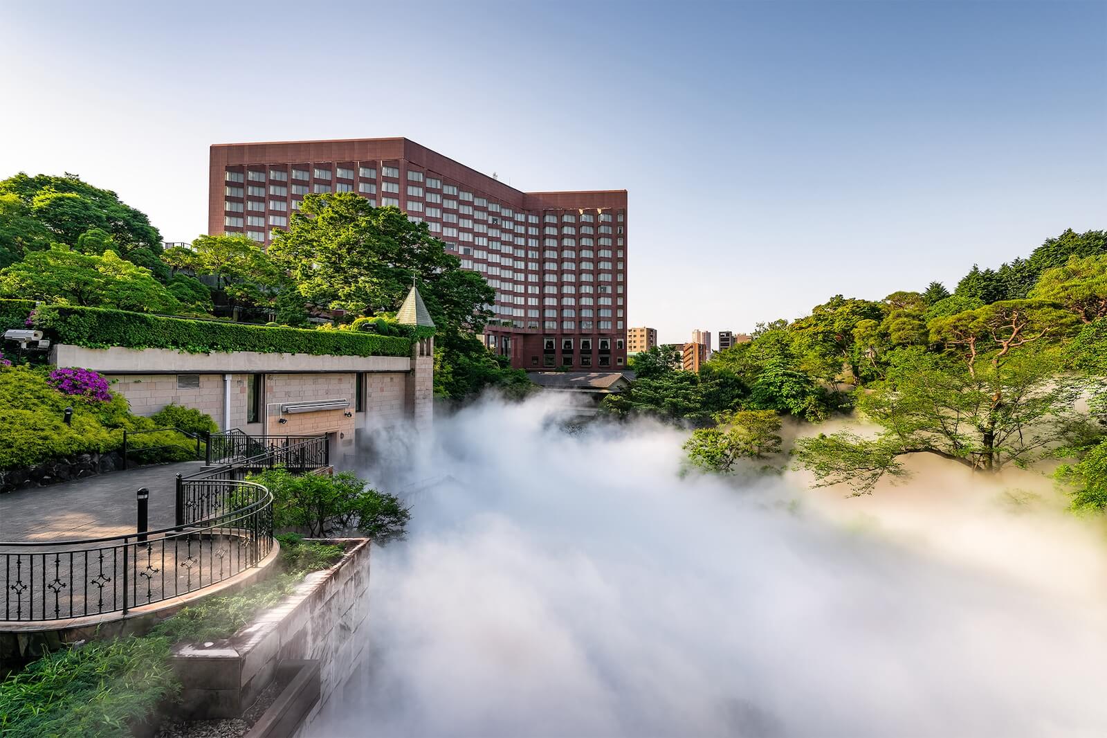 霧が立ち込める庭園越しに、背後に赤茶色のホテル建物が見える景観。
