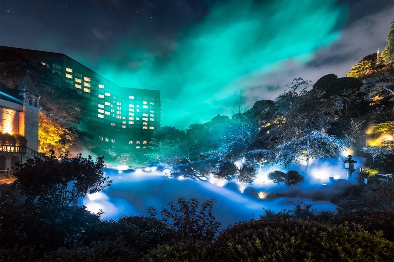夜の庭園にライトアップと霧の演出が広がり、背後のホテルとともに幻想的な光景が広がる風景。