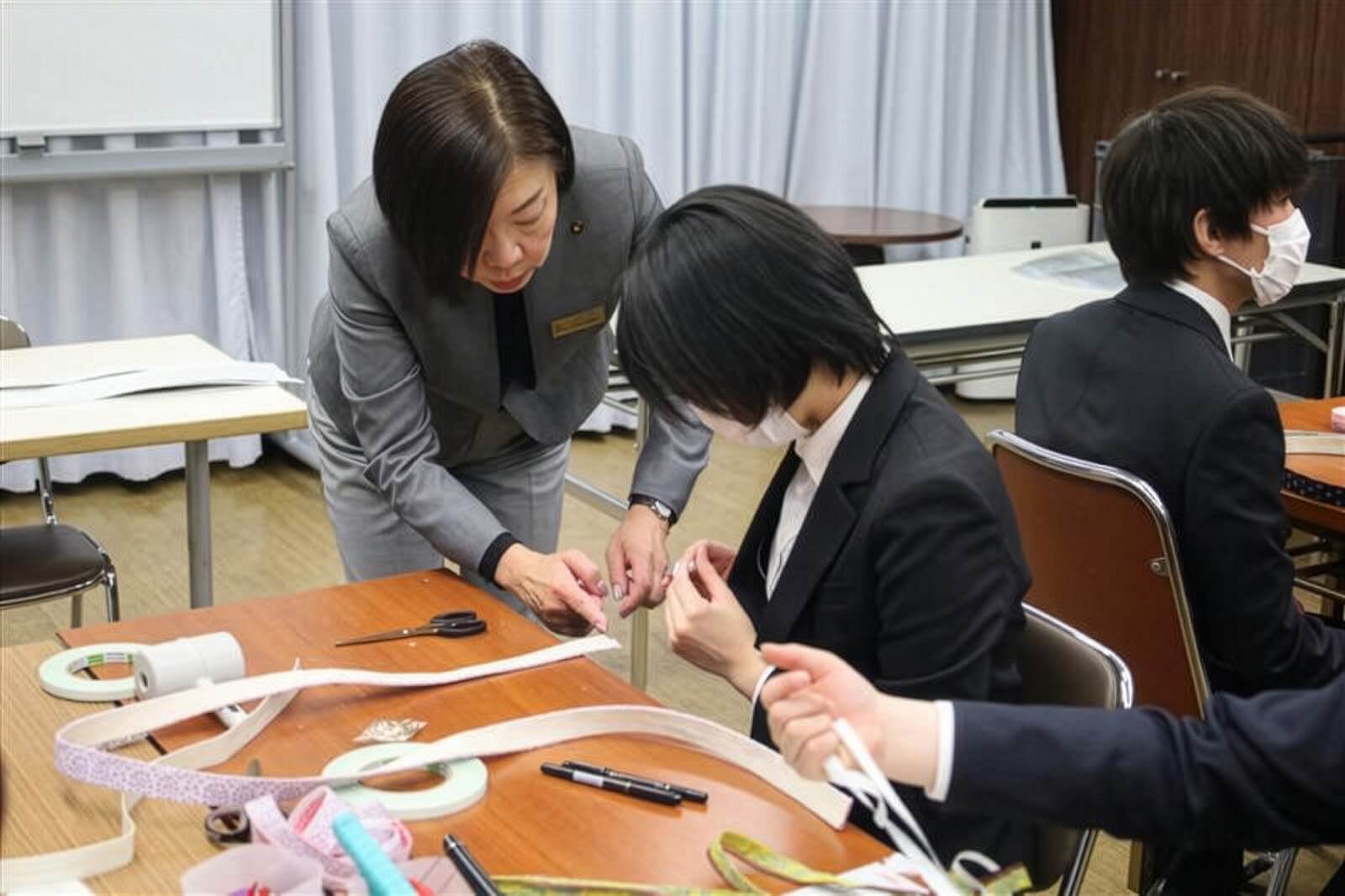 講師が受講者にリボン状の材料の扱い方を指導しながら、手元の作業をサポートしている様子。