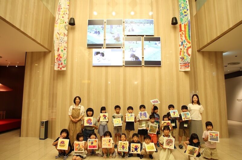 館内の吹き抜けロビーで、子どもたちが制作した作品を手に記念撮影している様子。
