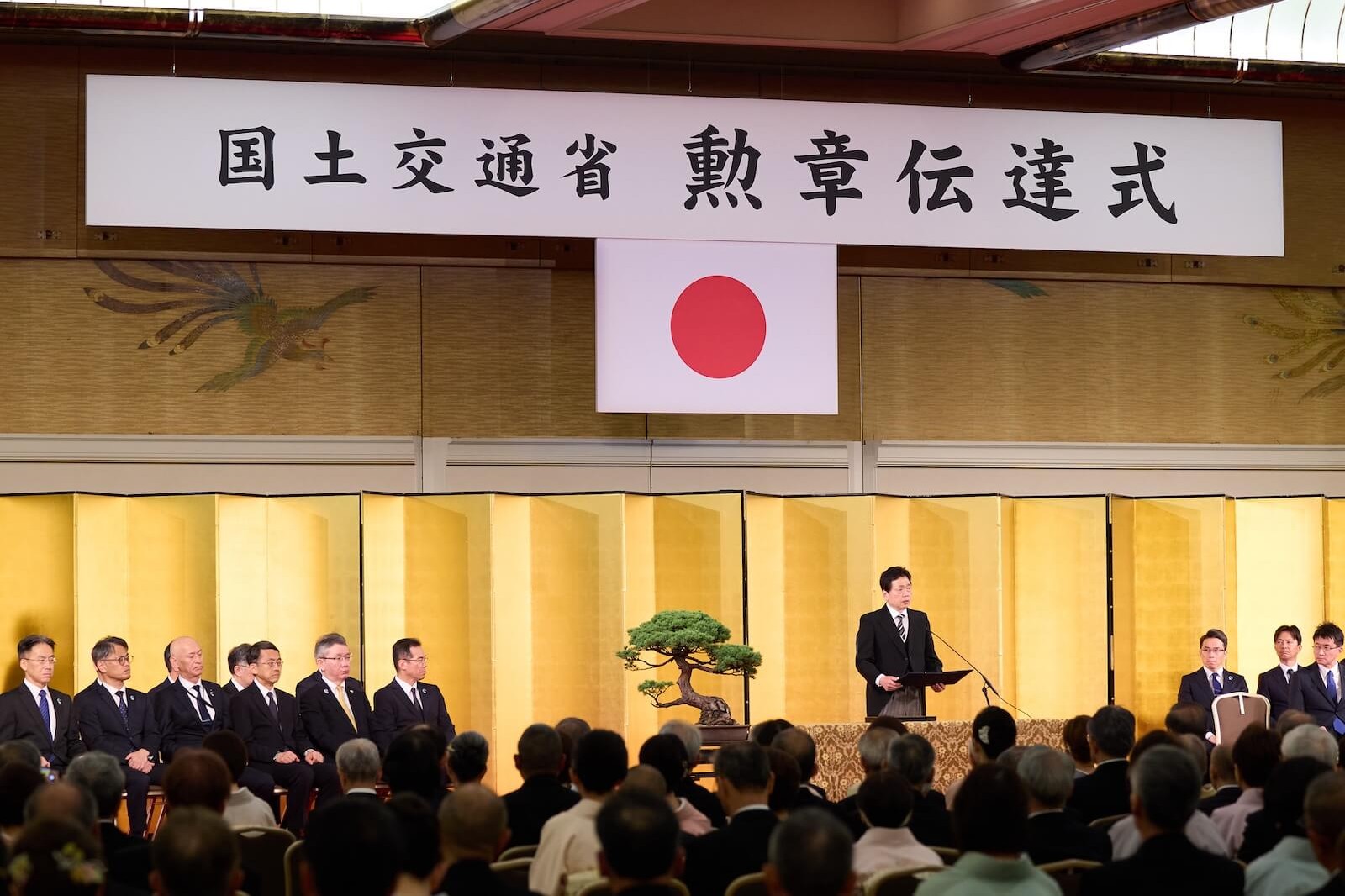 「国土交通省 勲章伝達式」の看板と日の丸が掲げられた会場で、壇上の登壇者が祝辞を述べる様子。金屏風を背景に、多数の出席者が式典に参加している。