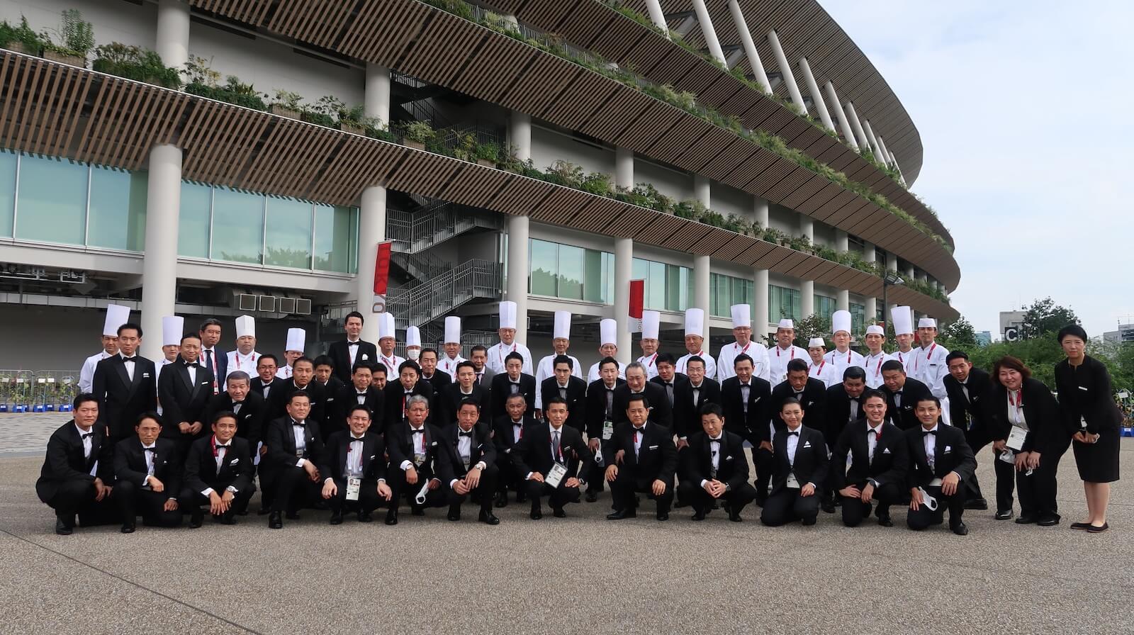 東京2020オリンピック・パラリンピック会場前で記念撮影を行うホテルスタッフとシェフの集合写真。タキシード姿やコックコート姿の参加者が並んでいる。