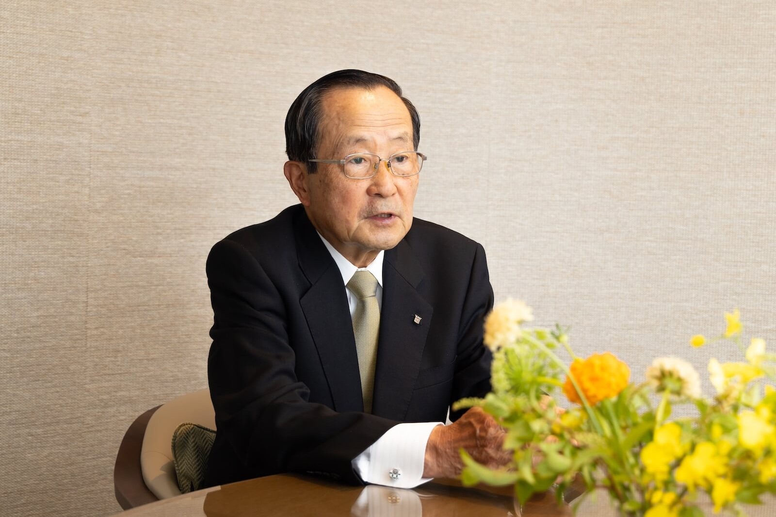 株式会社パレスホテル代表取締役会長の小林　節（こばやし・たかし）氏がインタビューに応じる様子。テーブル越しに着席し、手前には黄色や白の花が活けられた花瓶が置かれている。