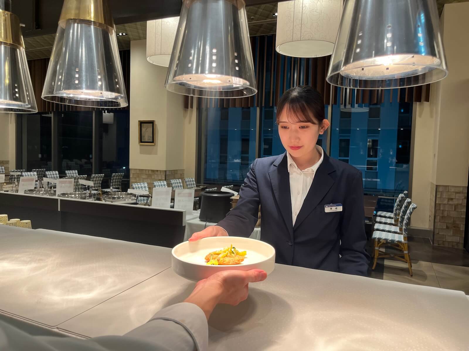 栗崎かれんさんが料理をサーブする様子。店内には大型のペンダントライトが並び、落ち着いた雰囲気のダイニング空間が広がる。