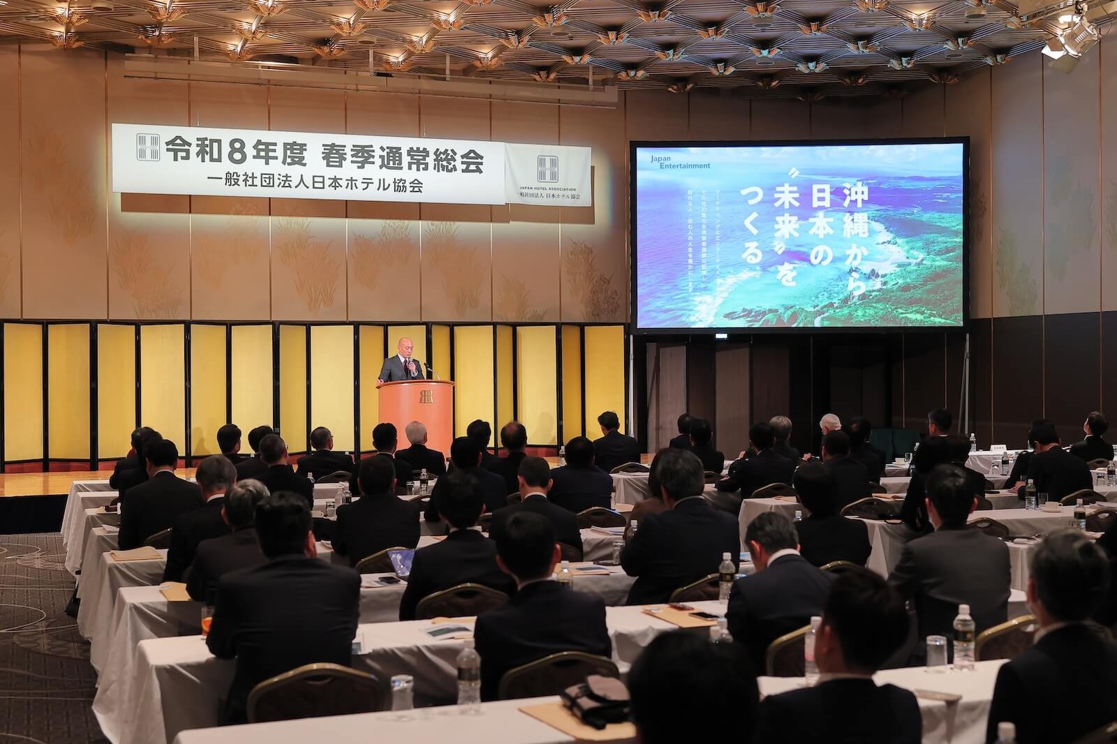 令和8年度春季通常総会の会場で、一般社団法人日本ホテル協会の総会が開催され、演壇での講演を参加者が着席して聴講している様子。スクリーンには「沖縄から日本の未来をつくる」の映像が映し出されている。