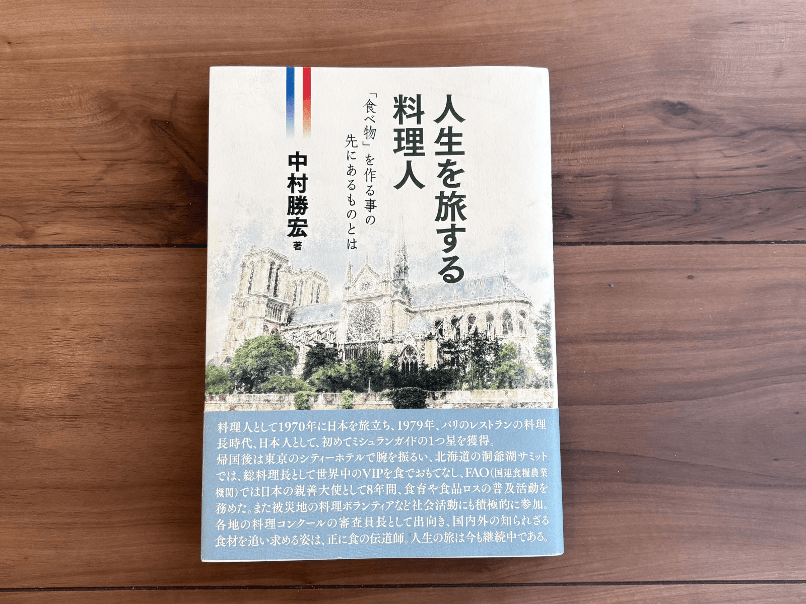 書籍表紙写真。表紙にはノートルダム大聖堂のイラストとともに、「人生を旅する料理人」「『食べ物』を作る事の先にあるものとは」「中村勝宏 著」と記載されている。下部には著者プロフィールが記載された帯があしらわれ、「料理人として1970年に日本を旅立ち、1979年、パリのレストランの料理長時代、日本人として、初めてミシュランガイドの1つ星を獲得。帰国後は東京のシティーホテルで腕を振るい、北海道の洞爺湖サミットでは、総料理長として世界中のVIPを食でおもてなし、FAO（国際連合食糧農業機関）では日本の親善大使として8年間、食育や食品ロスの普及活動を務めた。また被災地の料理ボランティアなど社会活動にも積極的に参加。各地の料理コンクールの審査員長として出向き、国内外の知られざる食材を追い求める姿は、正に食の伝道師。人生の旅は今も継続中である。」と書かれている。
