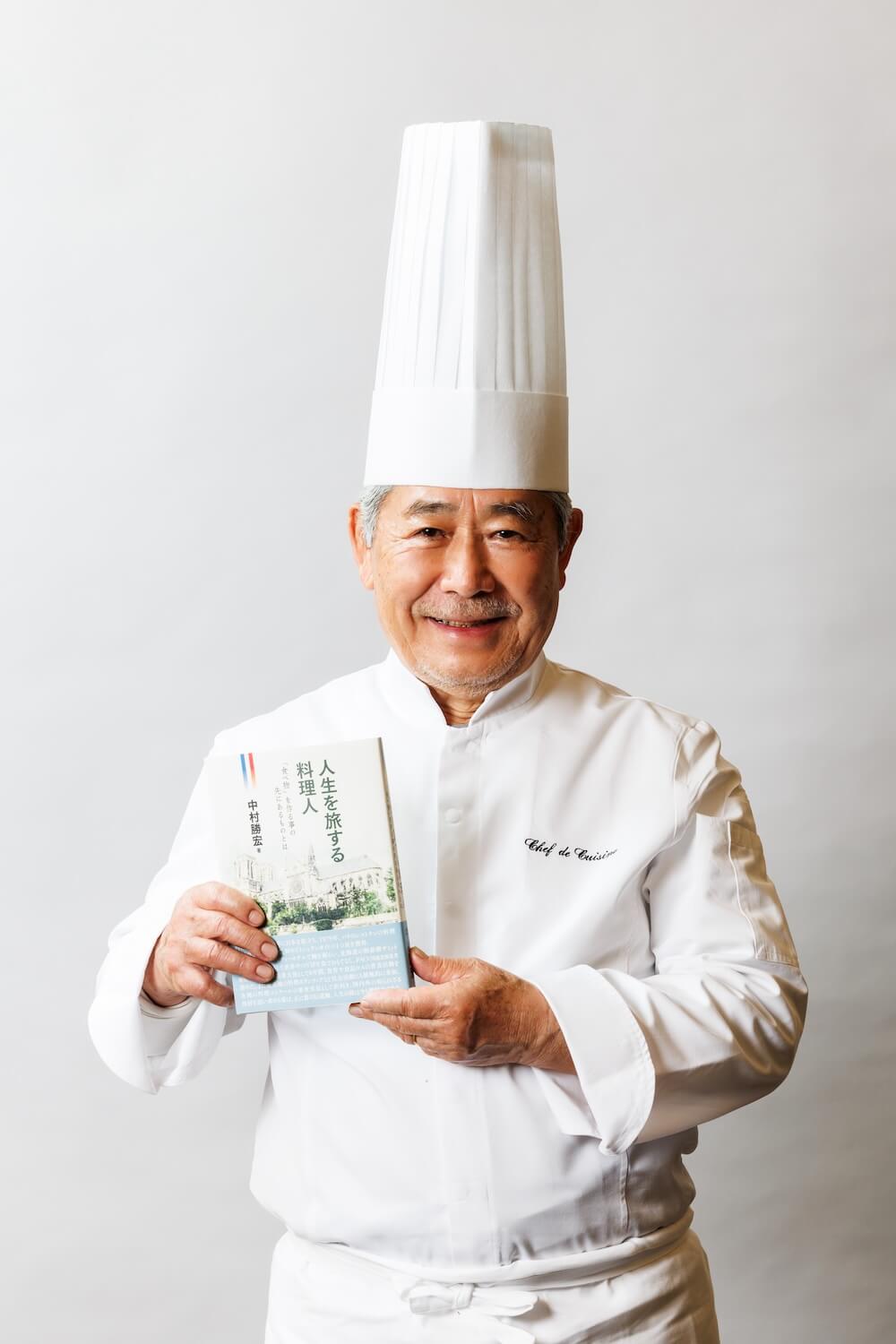 著書『人生を旅する料理人』を手にした中村勝宏シェフの肖像。白いコックコートとトック帽を着用し、笑顔で書籍を紹介している。