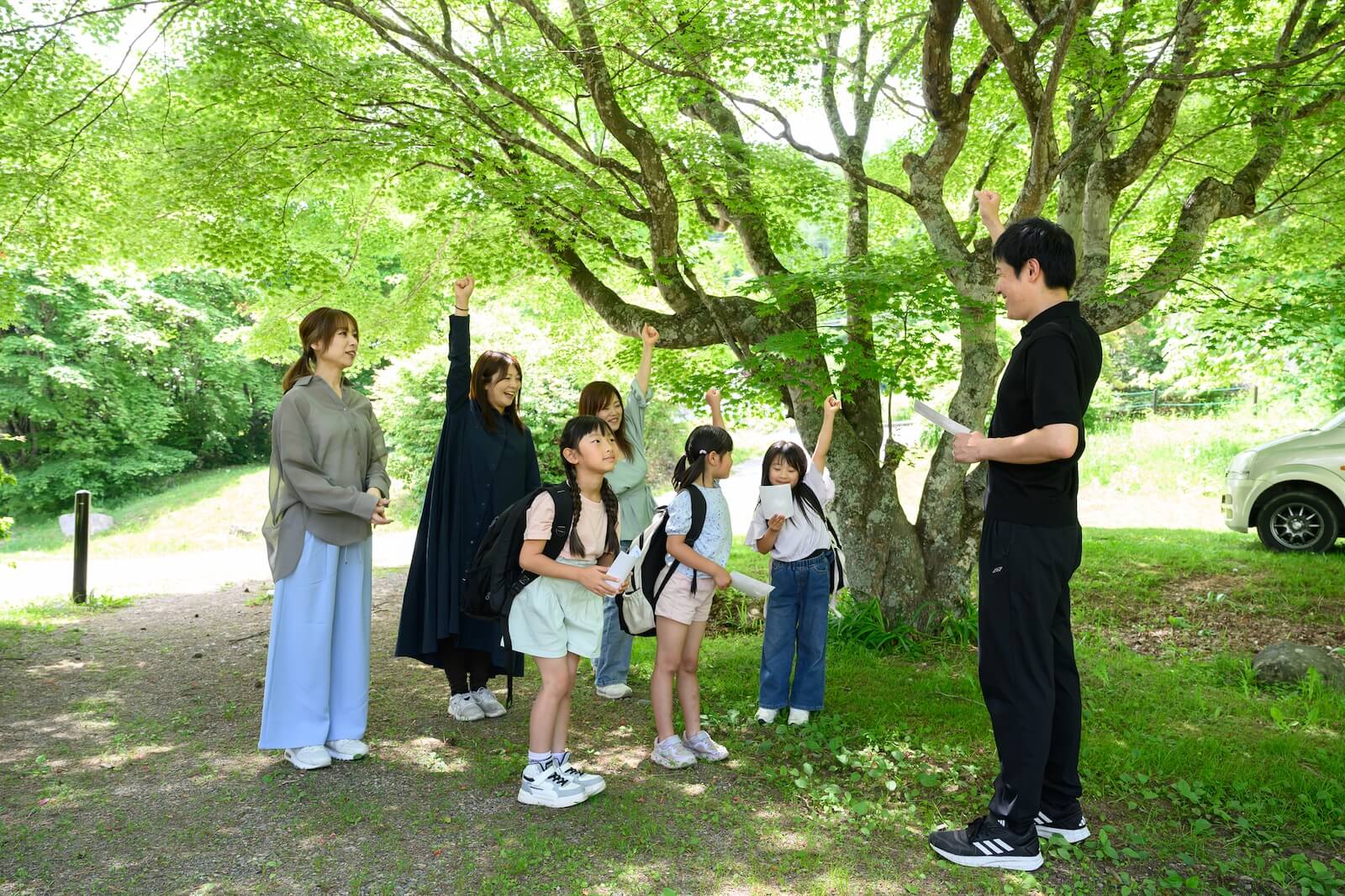 木陰で行われる自然体験イベントで、子どもたちや保護者らがスタッフの問いかけに手を挙げて答えている様子。