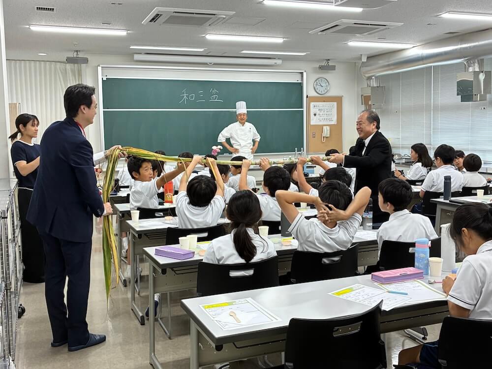 「和三盆」と書かれた黒板のある教室で行われた授業。小学生たちが席に着く中、大人らが長いサトウキビの両端を持ち、大人らに挟まれる形で小学生らもサトウキビを掴み上げている。白い調理服とコック帽を着た講師が教室前方から見守っている。