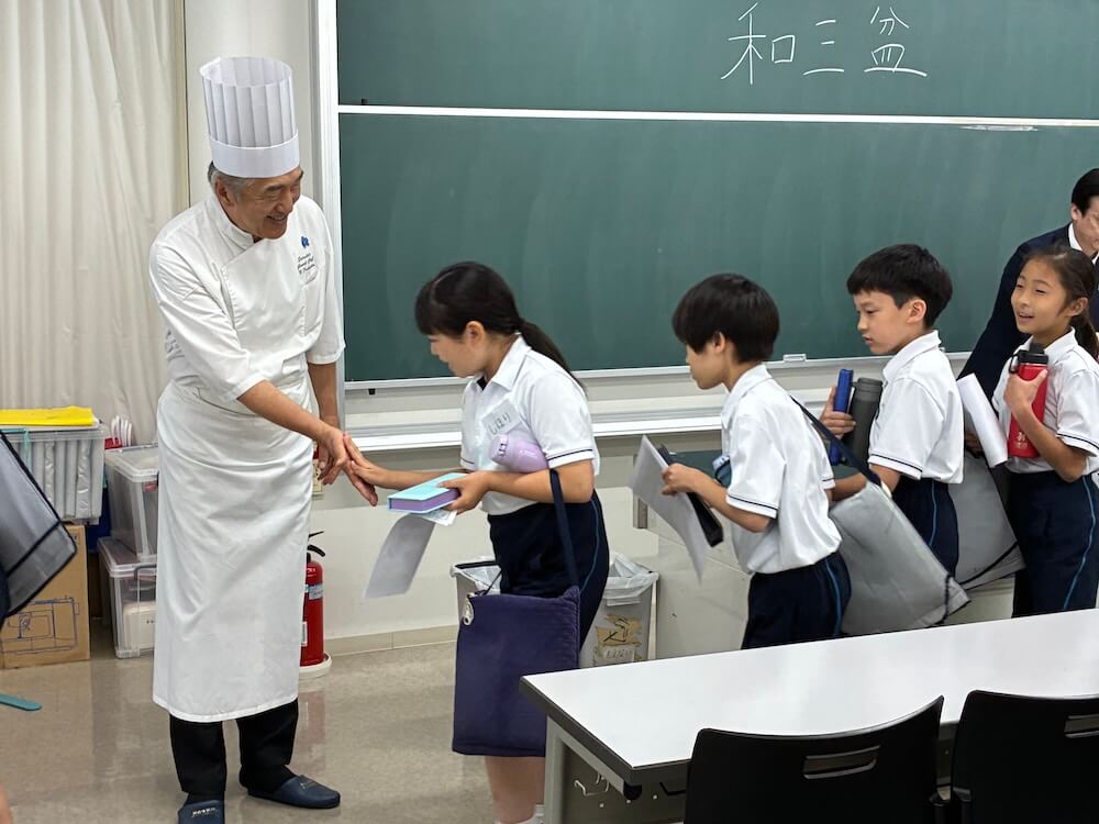 教室で白い調理服とコック帽を着た講師の前に小学生らが順に並び、講師の手のひらに小学生が一人ずつ自身の手で触れている様子。黒板には「和三盆」と書かれている。