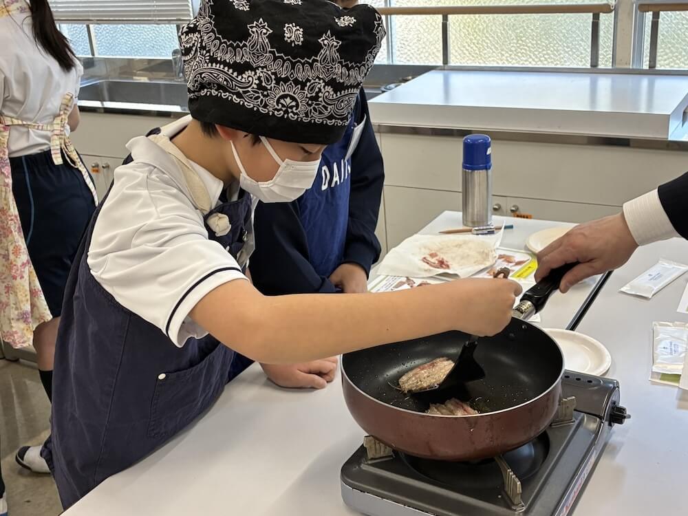調理実習で、バンダナとエプロン、マスクを着けた児童が、フライパンで魚を焼いている様子。大人にフライパンを支えてもらいながら、トングを使って調理している。