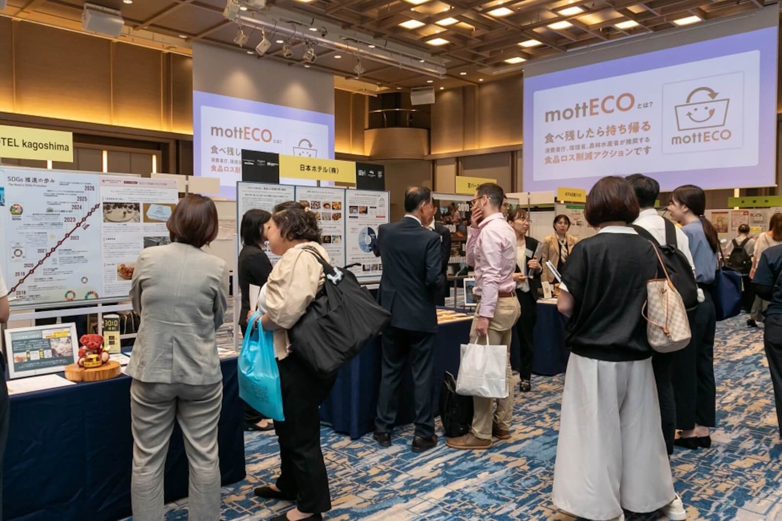 mottECOの取り組みを紹介する展示会場で、ホテル各社のパネル展示を見たり、担当者同士で情報交換したりする参加者。会場のスクリーンには「mottECOとは? 食べ残したら持ち帰る 食品ロス削減アクションです」と表示されている。
